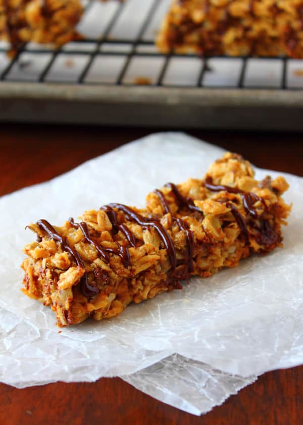 A Chewy Peanut Butter Granola Bar drizzled with chocolate and placed on a piece of parchment paper with others in the background on a cooling rack