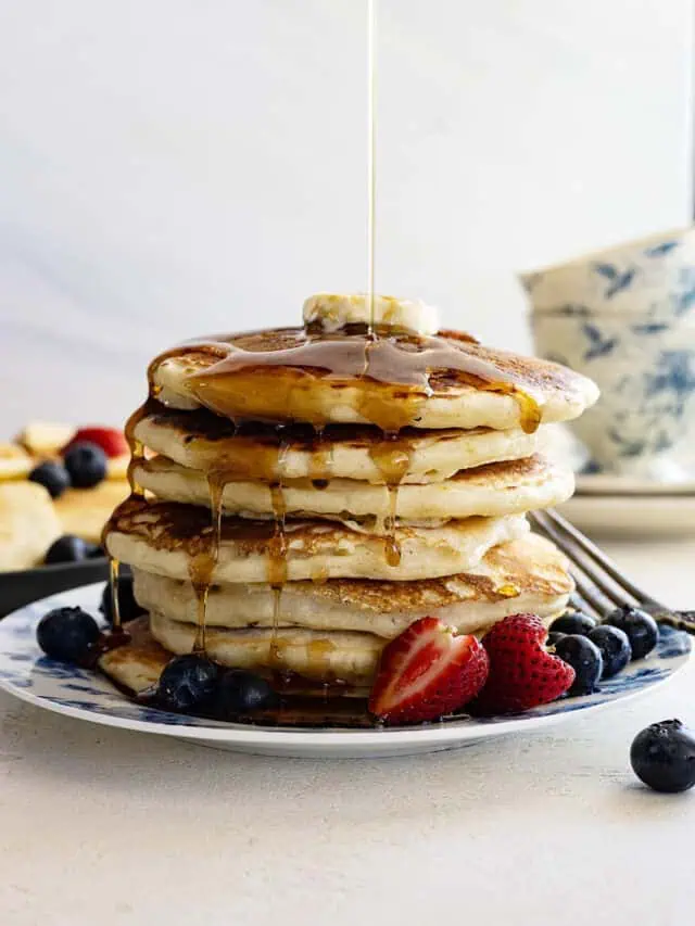 Tall stack of pancakes with butter on top and syrup being poured over the top.