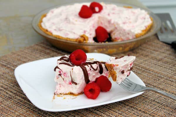 a slice of frozen raspberry cheesecake pie on a white plate with a forkful beside it, and the full pie in the background