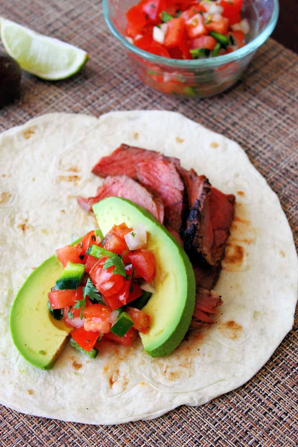a grilled steak tacos including a flour tortilla as a base, grilled steak, slices of avocado, and a bit of pico de gallo on top