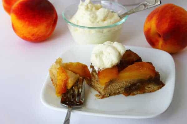 a slice of Peach Upside Down Cake on a white plate with a scoop of vanilla ice cream on top. A bowl of vanilla ice cream and a couple of whole peaches are in the background
