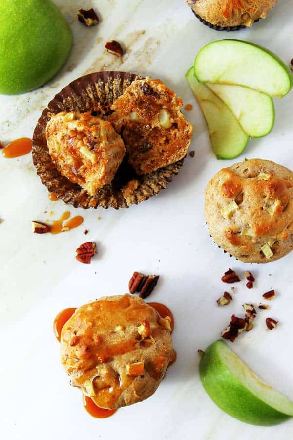 apple muffins with pecans on a white surface. one is cut open inside its muffin tin and drizzled with caramel. slices of granny smith apples and chopped pecans are scattered around