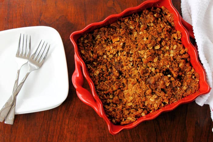 healthy apple crisp in a red square baking dish beside an empty white plate with two forks on it