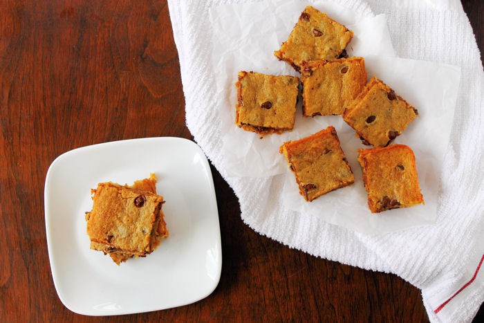 peanut butter blondes with banana and chocolate chips mixed in cut into squares. six blondies are on a white tea towel on the right and a few are stacked to the left on a white plate