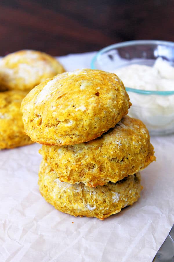three pumpkin biscuits stacked on top of each other in the foreground with more biscuits and a glass bowl full of maple butter in the background
