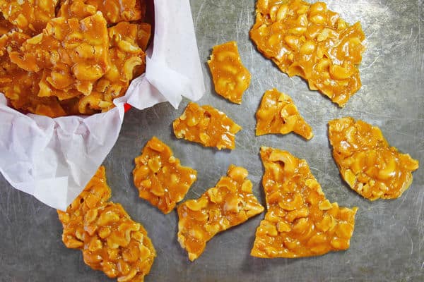 cashew brittle in a parchment lined bowl on the top left corner of the photo with different sized pieces of the brittle scattered on a grey surface