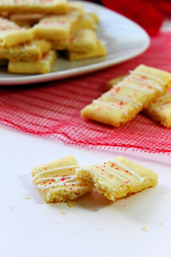 Swedish Butter Cookies on a white plate and out of focus in the background with a couple in the midground on a red fabric, and one cookie broken in half in the foreground on a white surface