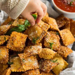 Toasted ravioli sprinkled with parmesan cheese and chopped basil. A child's hand is taking one to eat.