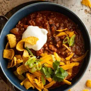 Overhead view of chili in a blue bowl topped with sour cream, cheese, and corn chips.