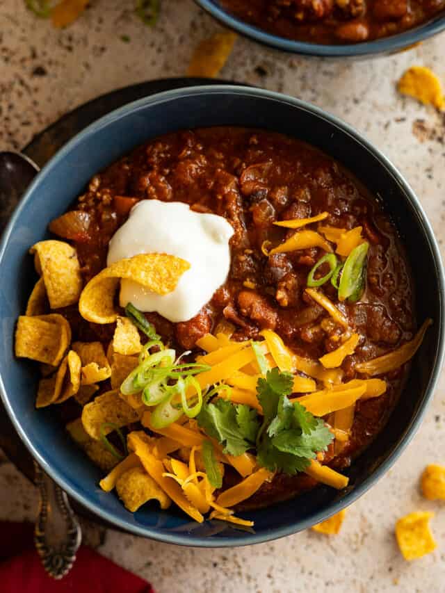 Overhead view of chili in a blue bowl topped with sour cream, cheese, and corn chips.