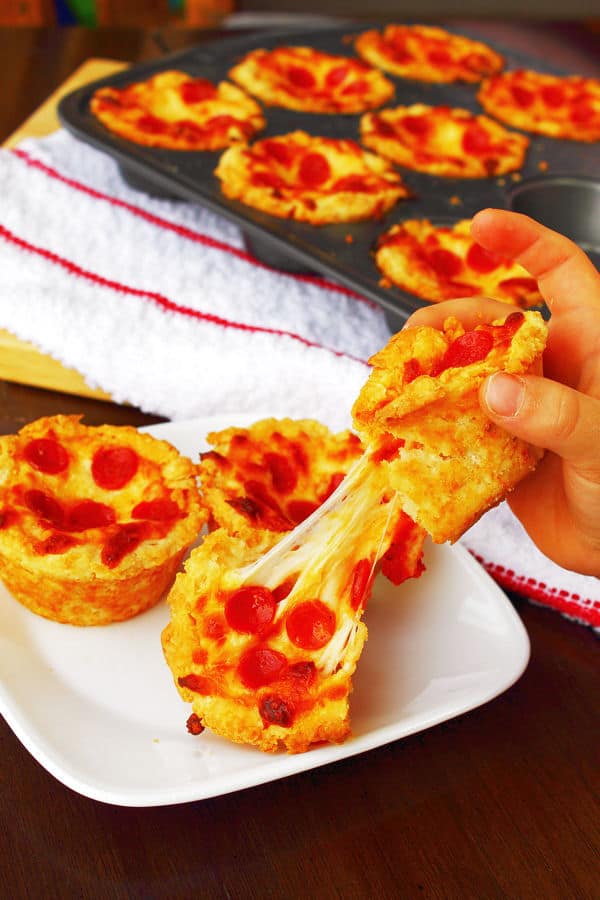 Mini Deep Dish Pizzas in a muffin tin in the background, and some on a white plate in the foreground with a hand pulling one of them apart with the cheese stretching from both halves