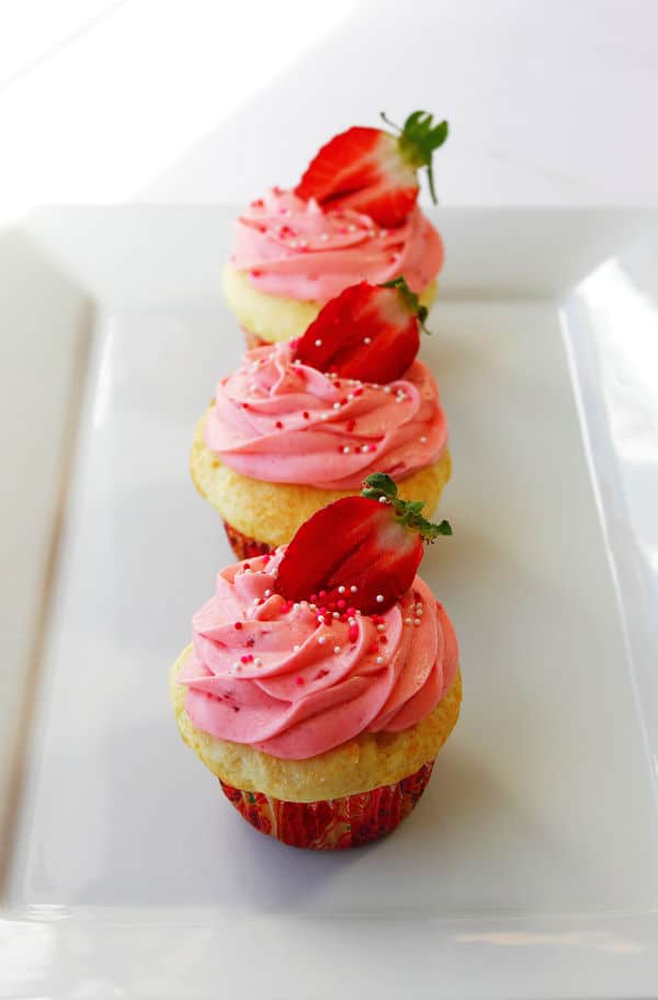 three Strawberry Shortcake Cupcakes in a line on a white serving platter with sprinkles and a halved strawberry on top
