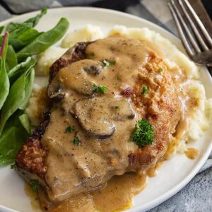 A pork chop on a plate with mash potatoes and topped with a creamy mushroom gravy.