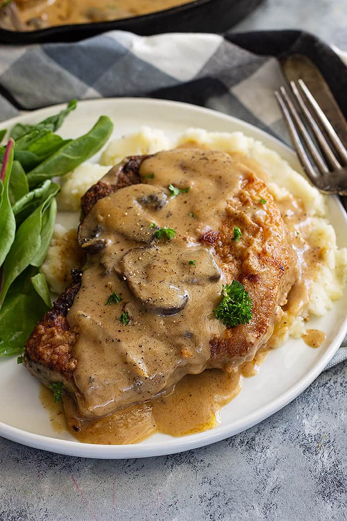 A pork chop on a plate with mash potatoes and topped with a creamy mushroom gravy. 