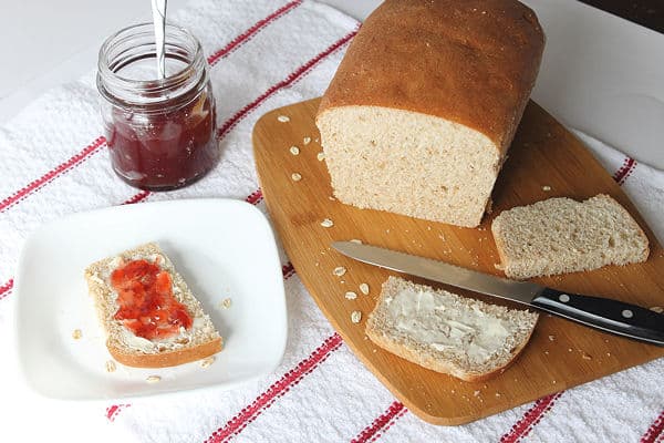Whole Wheat Oatmeal Bread on a cutting board with a few slices around. one is on a plate with butter and jam, and a jam jar sitting next to it