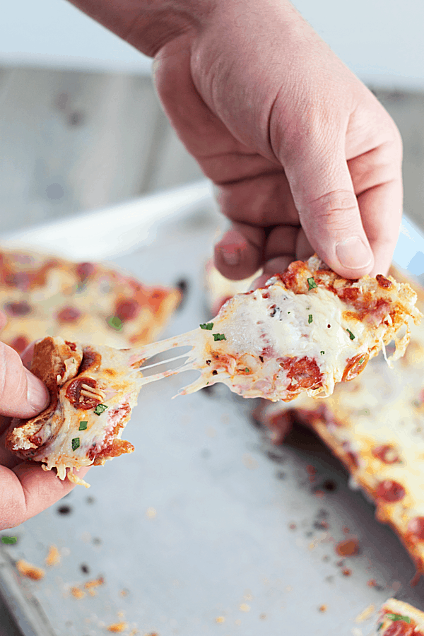 hands pulling a piece of this homemade french bread pizza recipe apart with the cheese stringing between both pieces and the remaining pizza on a baking sheet in the background