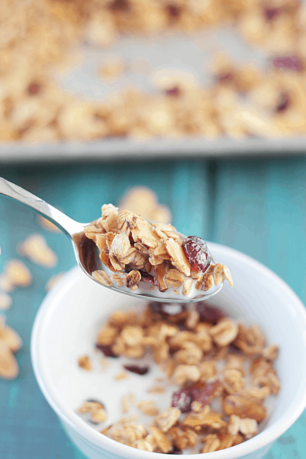 a scoop of homemade granola with maple and almond on a spoon with a bowl of granola and milk in the background
