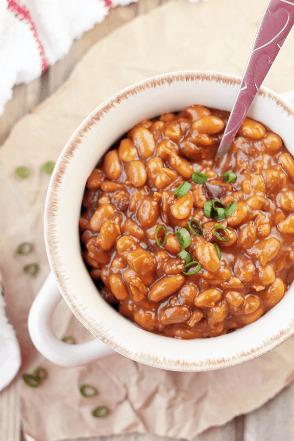 an overhead shot of baked beans in a white bowl with a spoon sticking out of it, and chives garnishing the top