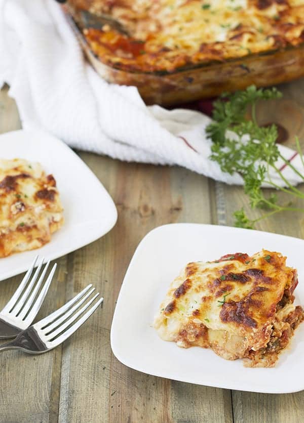 squares of lasagna cut from the larger casserole dish in the background on white plates with two forks sitting between the plates
