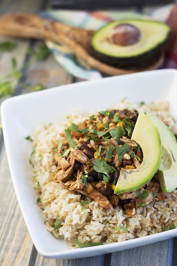 a white bowl of cooked rice with a scoop of slow cooker chicken on top garnished with avocado and herb garnish