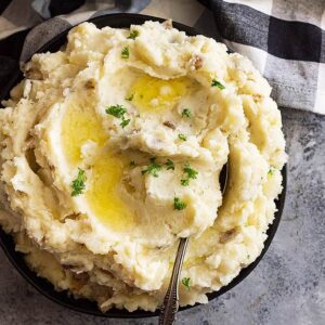 Large bowl of slow cooker mashed potatoes with pools of melted butter and chopped parsley for garnish.