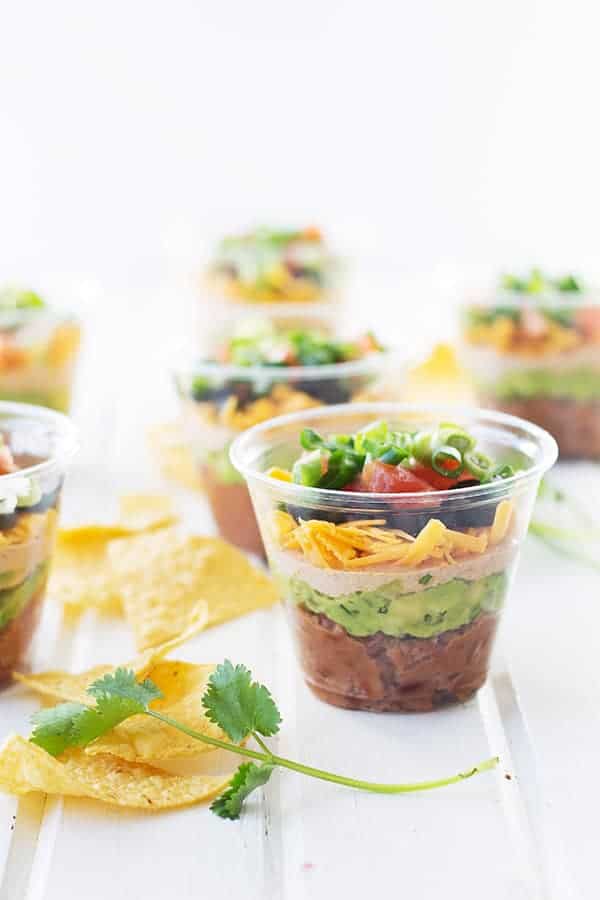 individual seven layer taco dip cups scattered around a table with tortilla chips around
