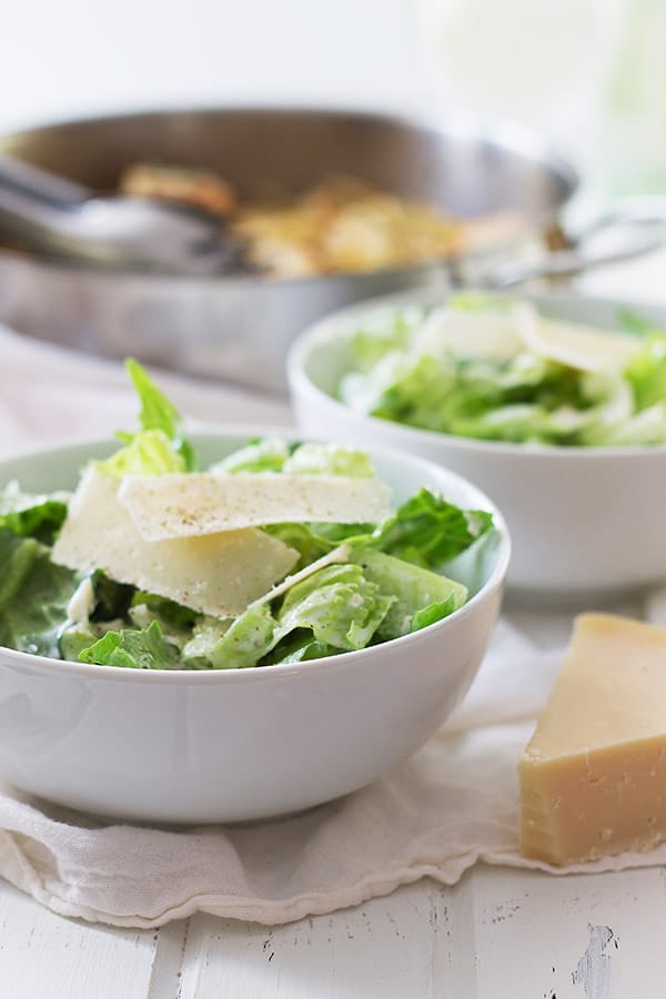 Close up of simple Caesar salad in a circular white bowl with a Parmesan wedge to the side. 