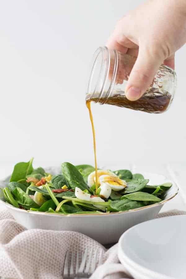 Side view of warm spinach salad with bacon with bacon dressing being poured from a mason jar above the salad.