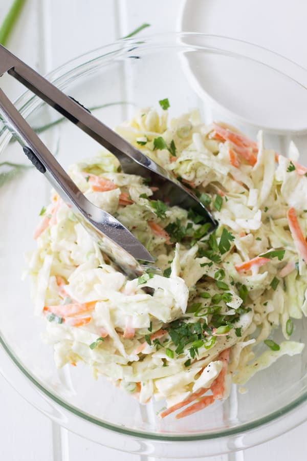 Shredded cabbage and carrot coleslaw in glass mixing bowl with tongs.