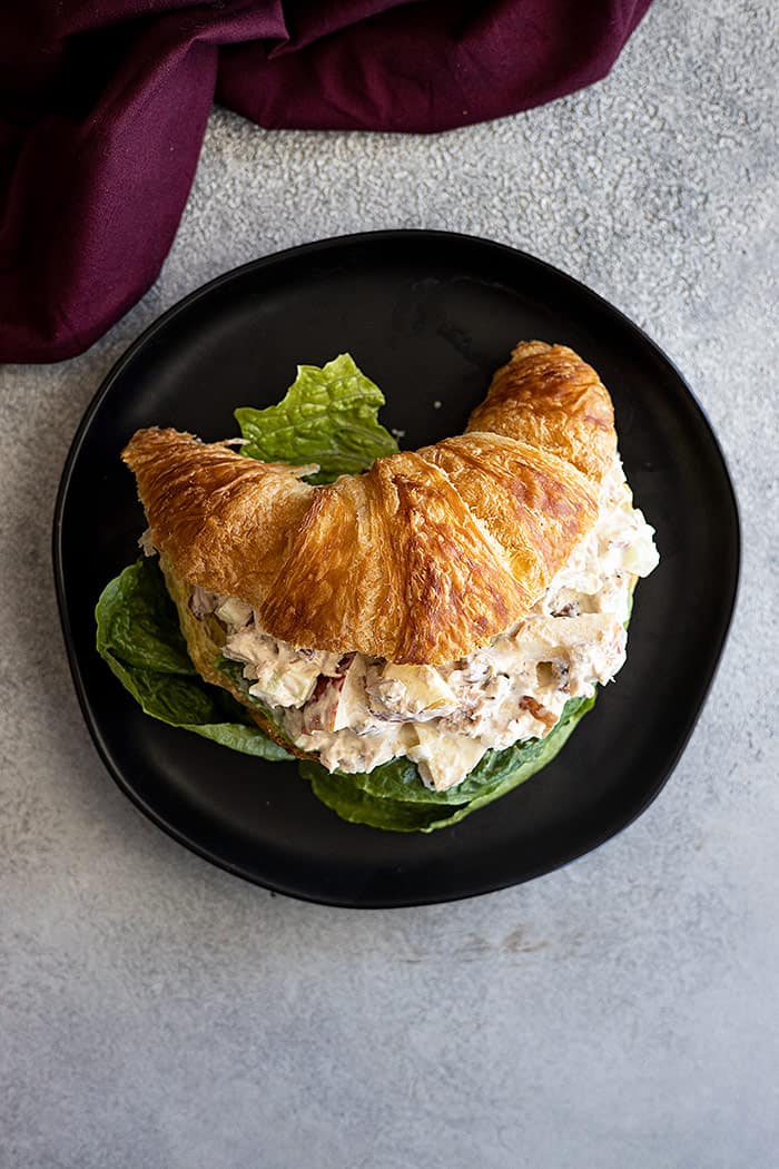 Overhead view of apple tuna salad on a croissant on a black plate with a lettuce leaf. 