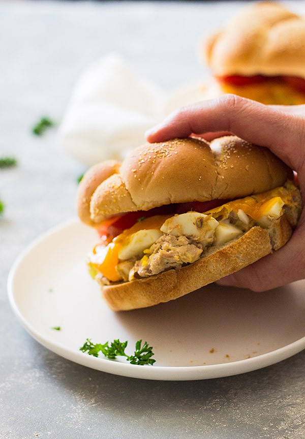 Hand lifting tuna melt sandwich on hamburger bun from white plate.