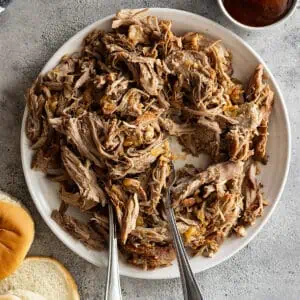 Overhead view of pulled pork on a white plate with two forks in the meat. Buns and bbq sauce off to the side.