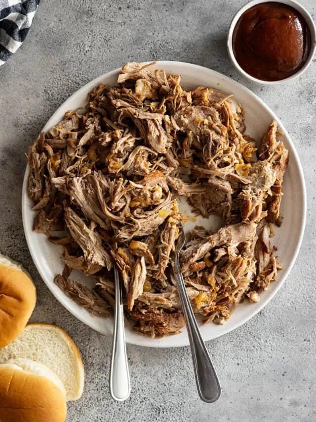 Overhead view of pulled pork on a white plate with two forks in the meat. Buns and bbq sauce off to the side.