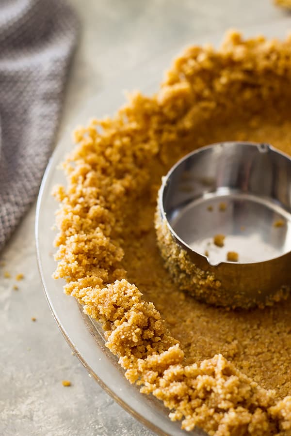 Graham cracker crust being pressed into pie pan with measuring cup. 