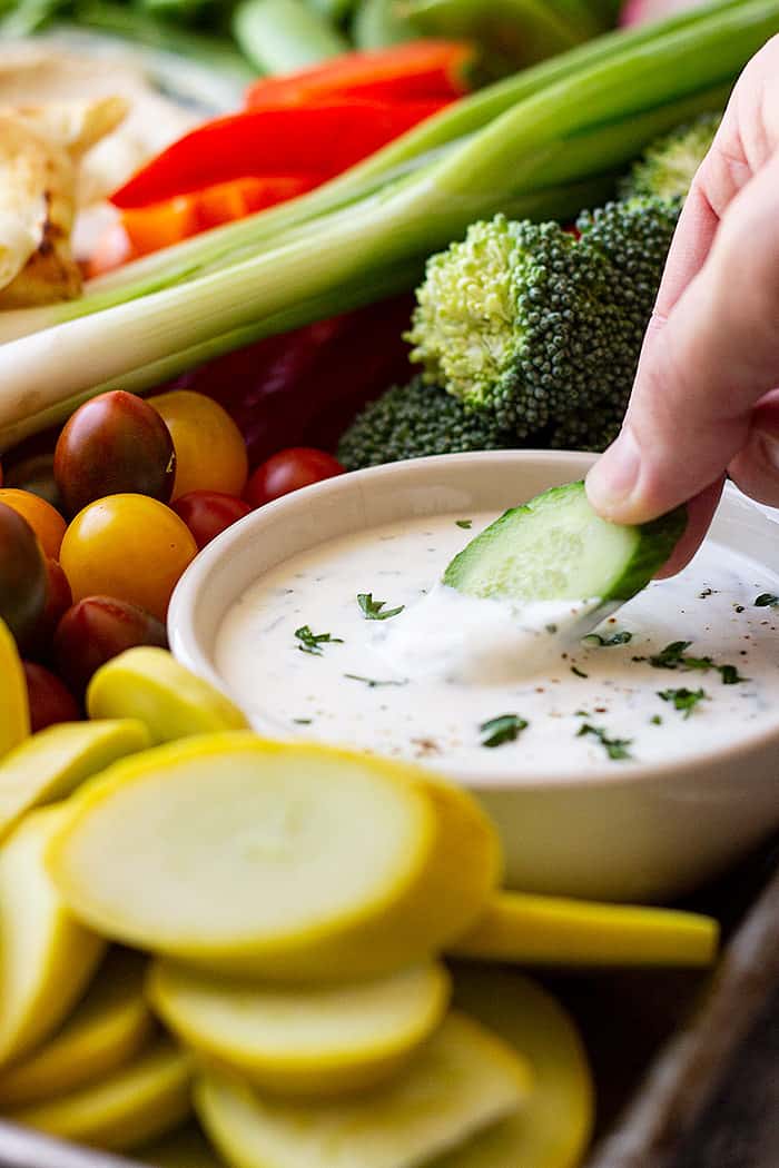 A hand with a cucumber dipping into a bowl of homemade ranch dressing. 
