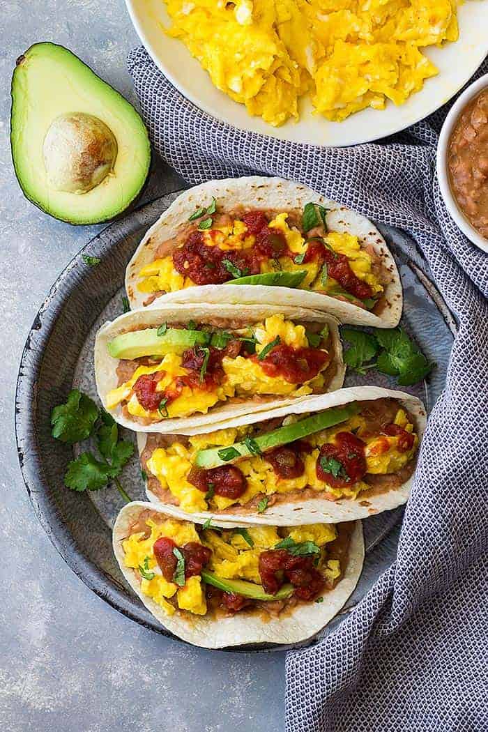 Bean and cheese breakfast tacos topped with salsa and avocado with shredded cheddar off to the side. 