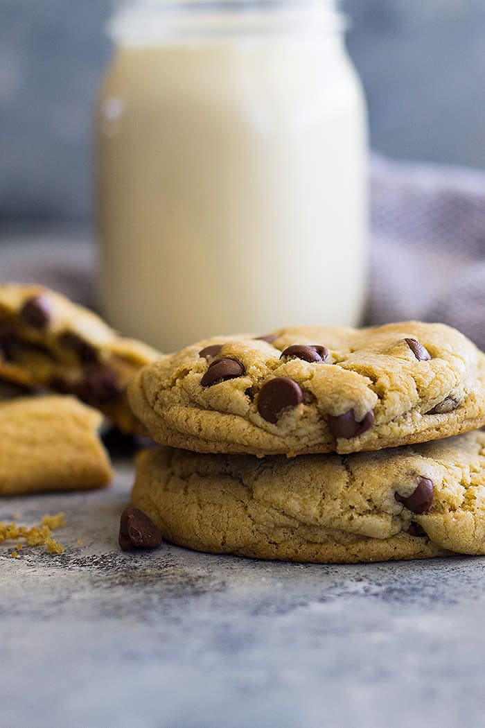 Two chocolate chip cookies stacked with milk and a broken cookie in the background. 