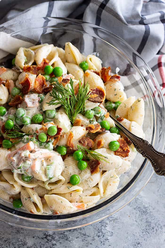 Pasta salad in a large bowl with a serving spoon in the salad. Garnished with crumbled bacon and peas.