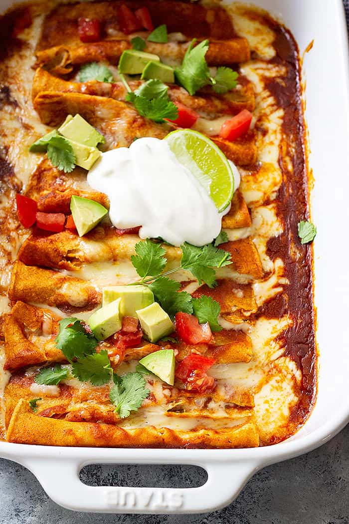 Cheese enchiladas in a white baking pan topped with avocado, tomatoes, and sour cream. 