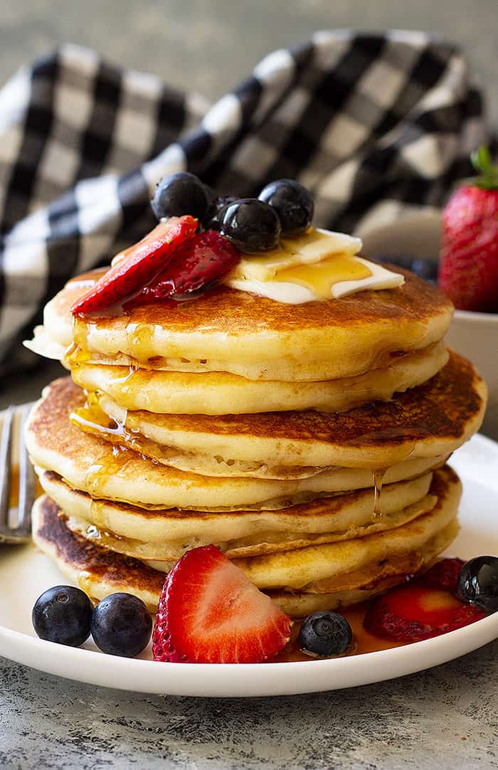 Tall stack of fluffy buttermilk pancakes dripping with maple syrup and topped with butter and berries. 