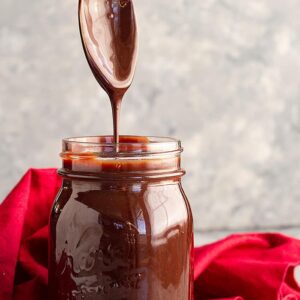 A spoon pulled straight up from a jar of homemade hot fudge sauce letting it drizzle back into the jar.