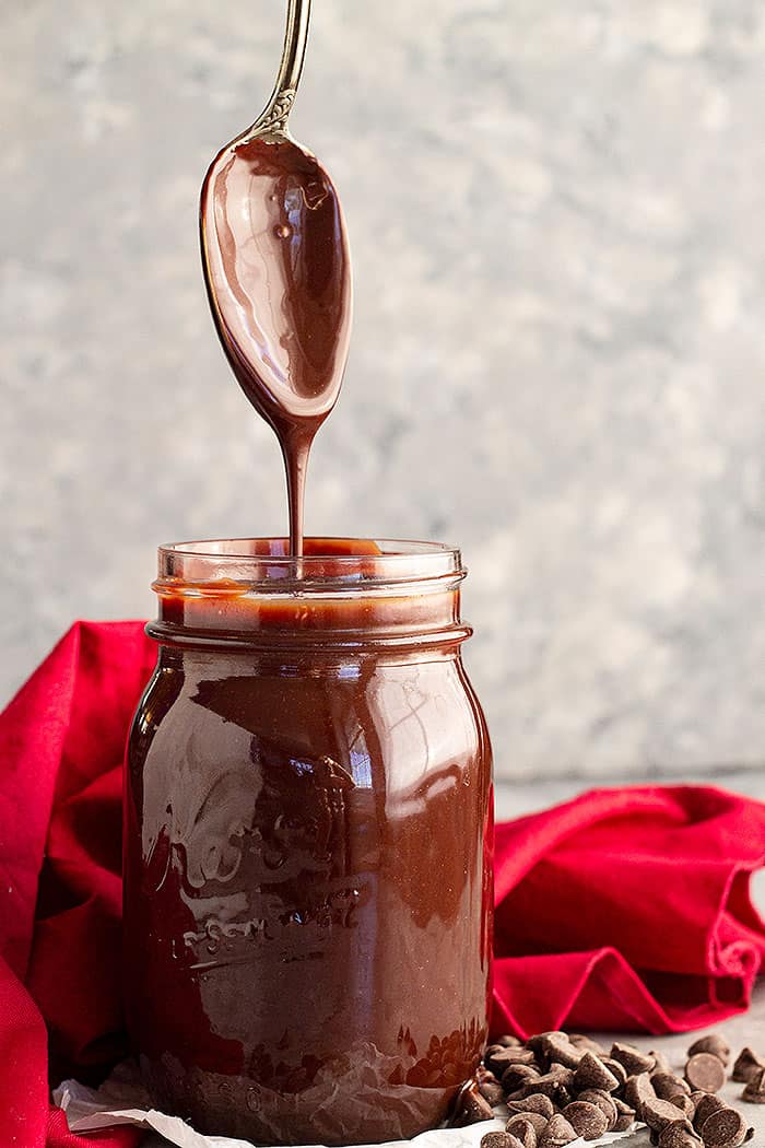 A spoon pulled straight up from a jar of homemade hot fudge sauce letting it drizzle back into the jar.