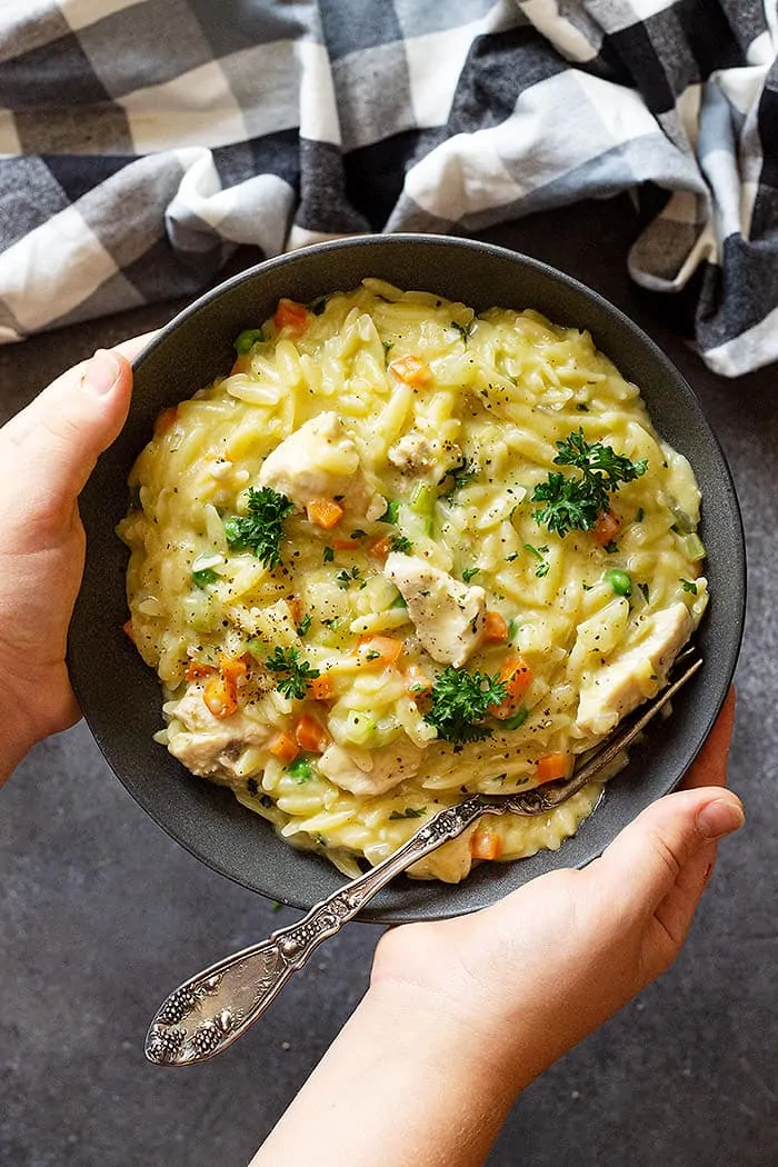 Hands holding a bowl of chicken orzo with a spoon in it.