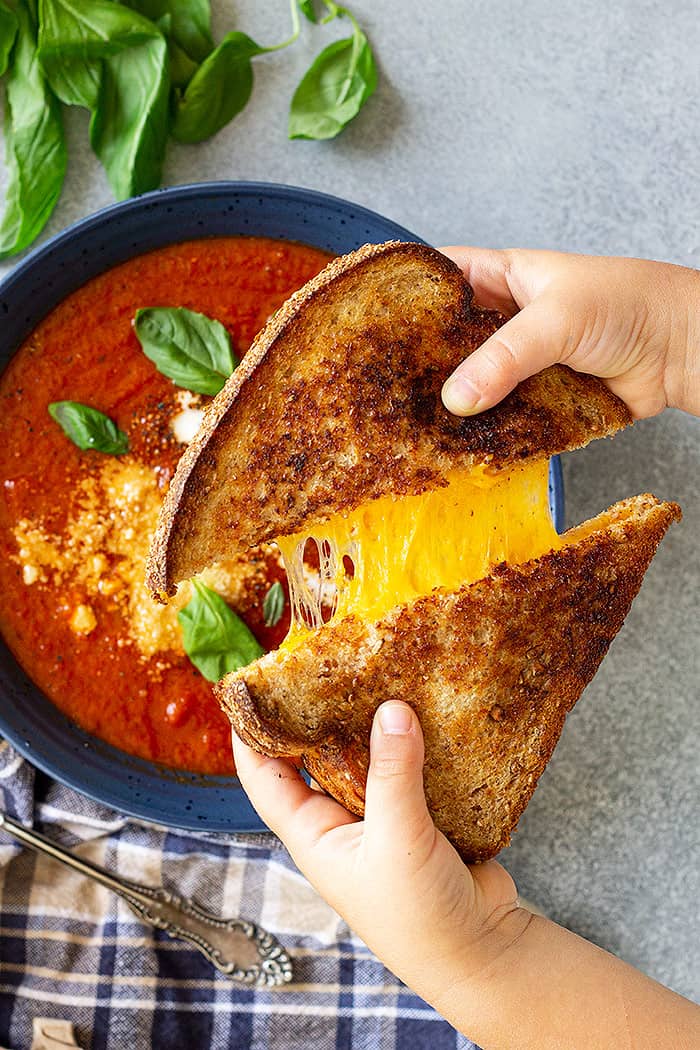 Hands pulling apart a grilled cheese sandwich over a bowl of soup.