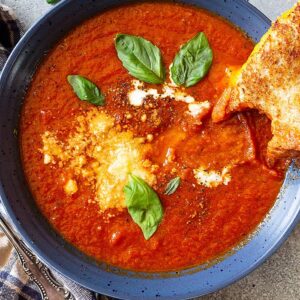 Top down view of a bowl of tomato soup garnished with basil, parmesan cheese, and swirl of cream, and a grilled cheese off to the side.