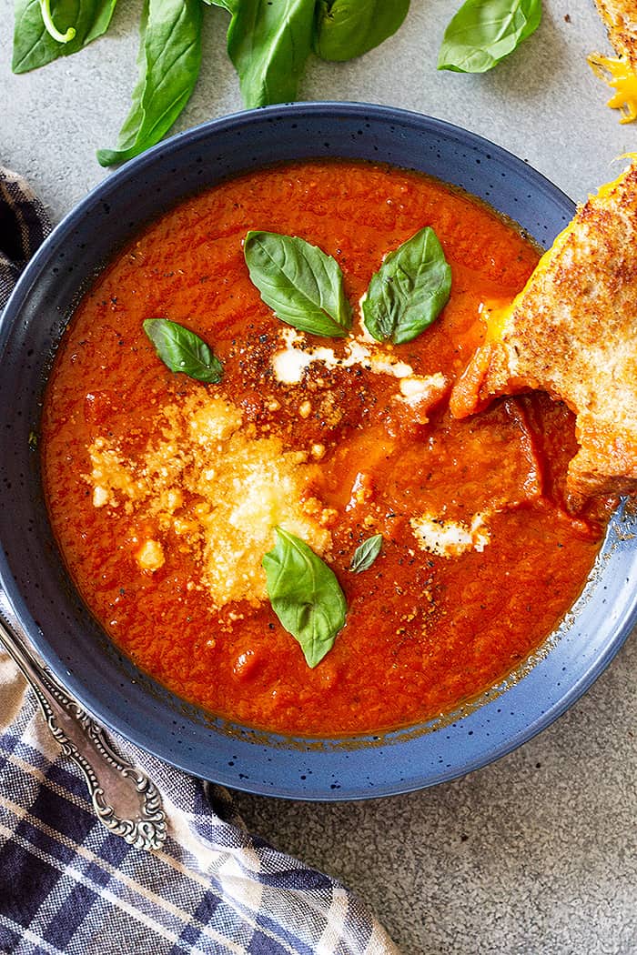 Top down view of a bowl of tomato soup garnished with basil, parmesan cheese, and swirl of cream, and a grilled cheese off to the side. 