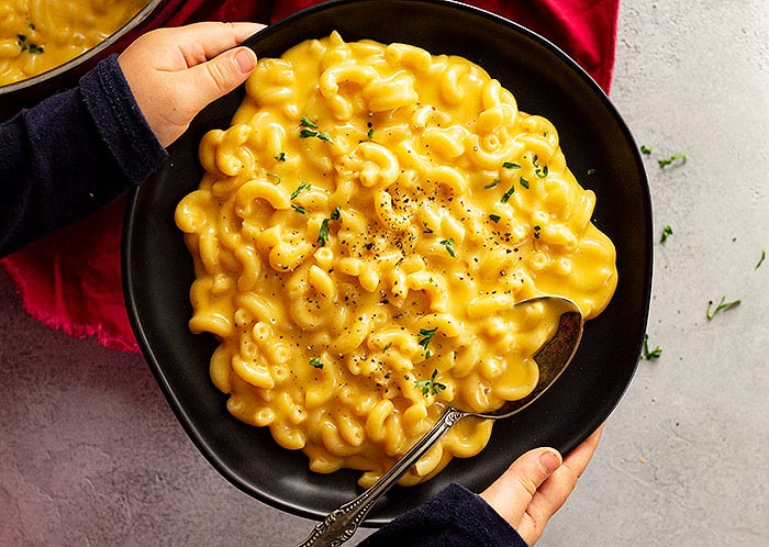 A child's hands holding a bowl of mac and cheese garnished with black pepper and parsley. 