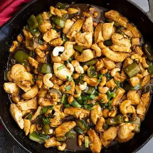 Overhead view of cashew chicken in a cast iron skillet garnished with green onions. Red napkin off to the side.