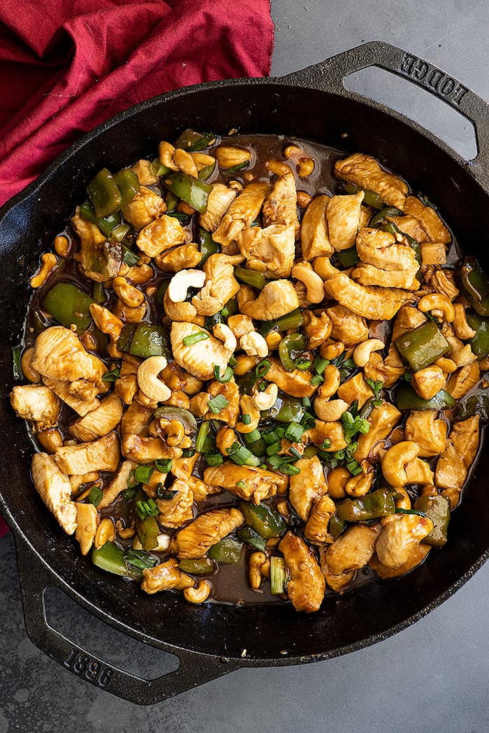Overhead view of cashew chicken in a cast iron skillet garnished with green onions. Red napkin off to the side.