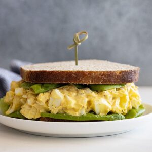 An egg salad sandwich on a plate. Sandwich also has a large lettuce leaf and a decorative toothpick in the center.